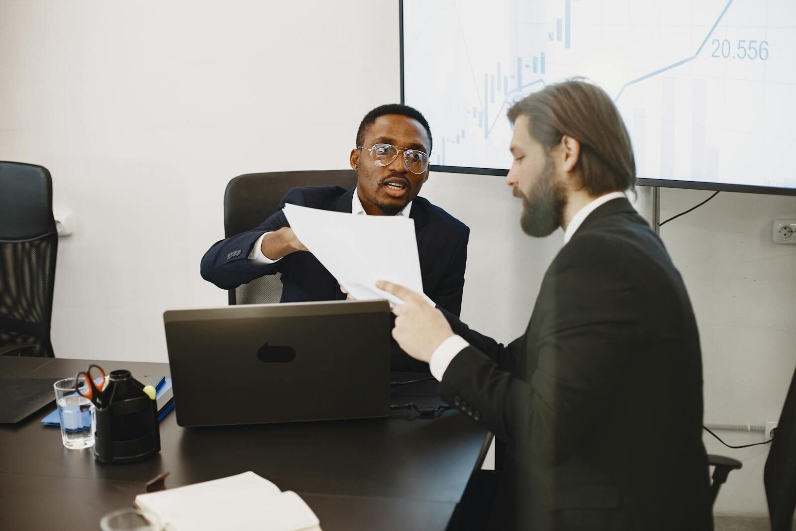 Two businessmen in suits discussing documents at an office meeting with a laptop and charts.