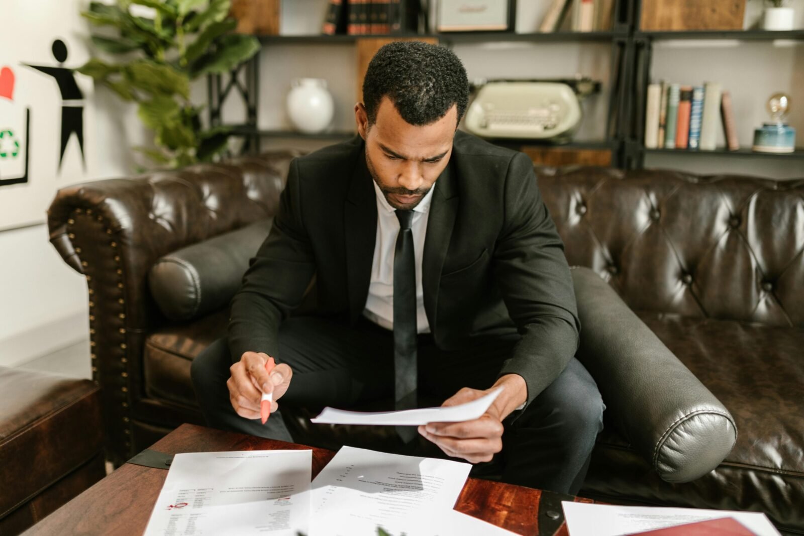 A businessman in a suit attentively reviews paperwork on a brown leather sofa.